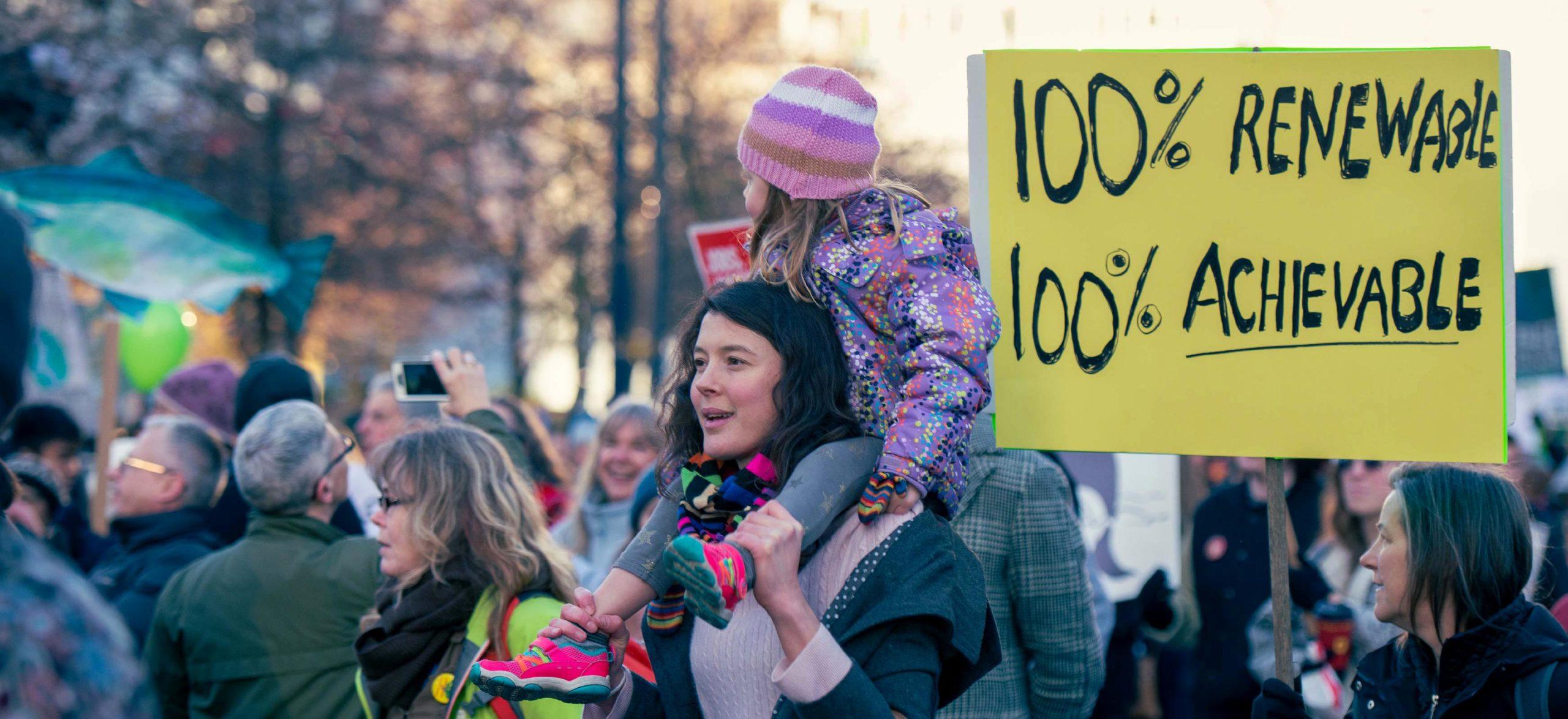 A group of people at a climate change protest. In the centre of the photo is a young woman with a child on her shoulders