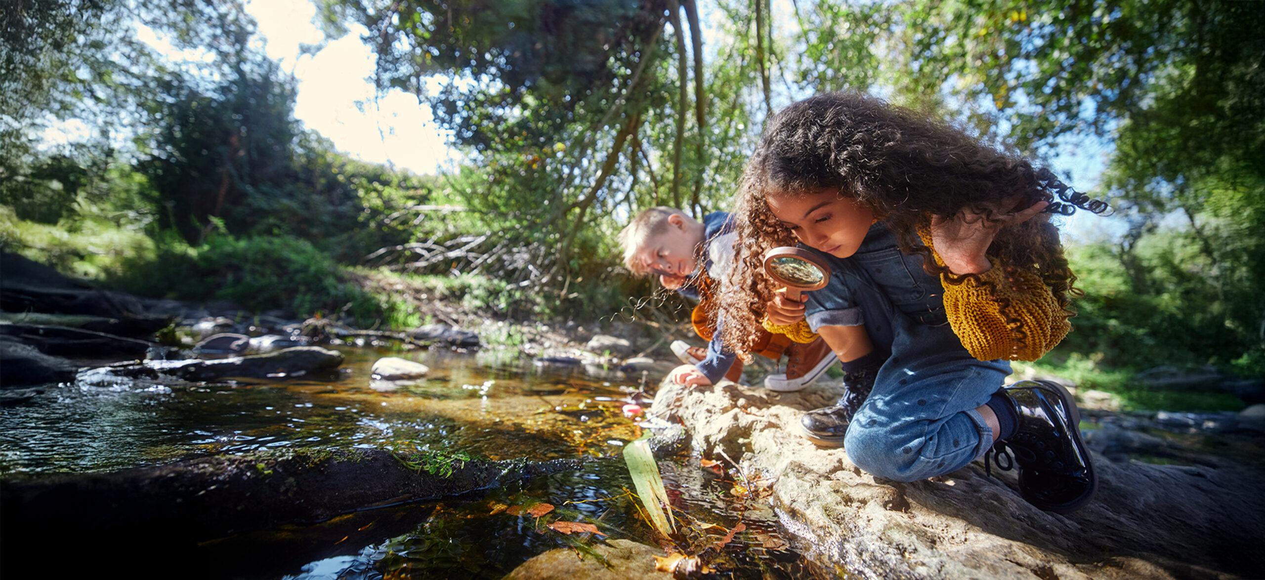 Two young people crouch down and look at river with magnifying glass.