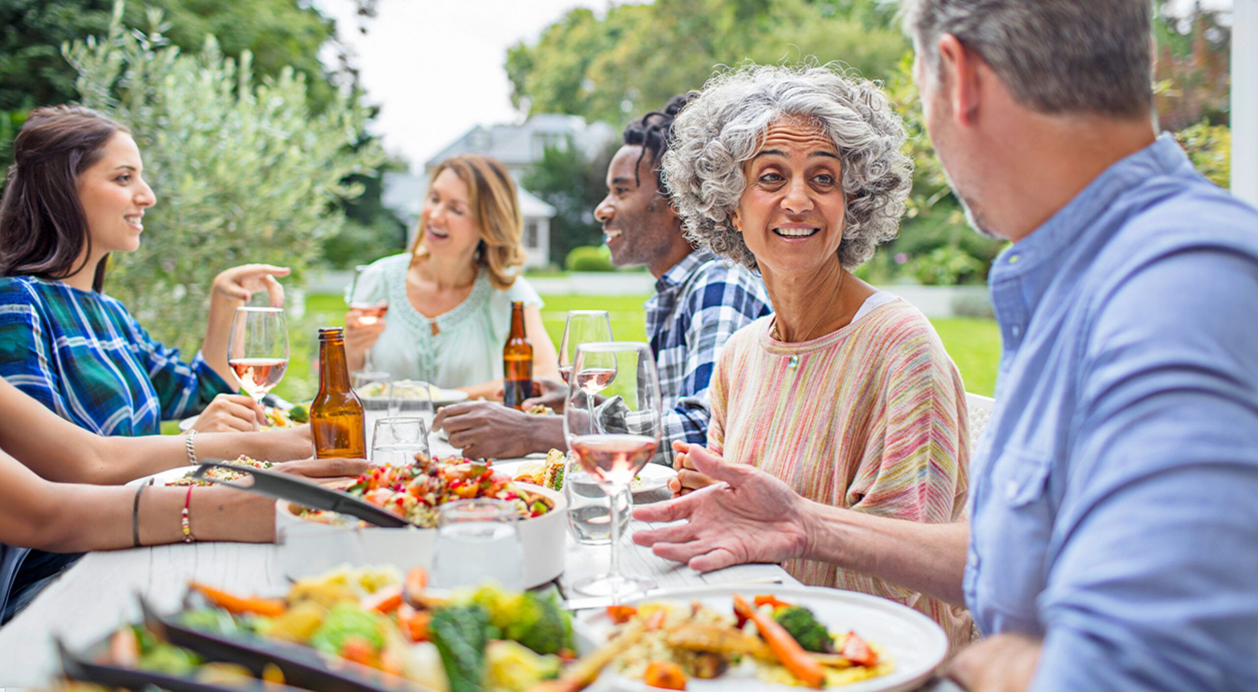 Mature friends talking while having lunch in backyard. Multi-ethnic males and females are enjoying garden party. They are wearing casuals.
