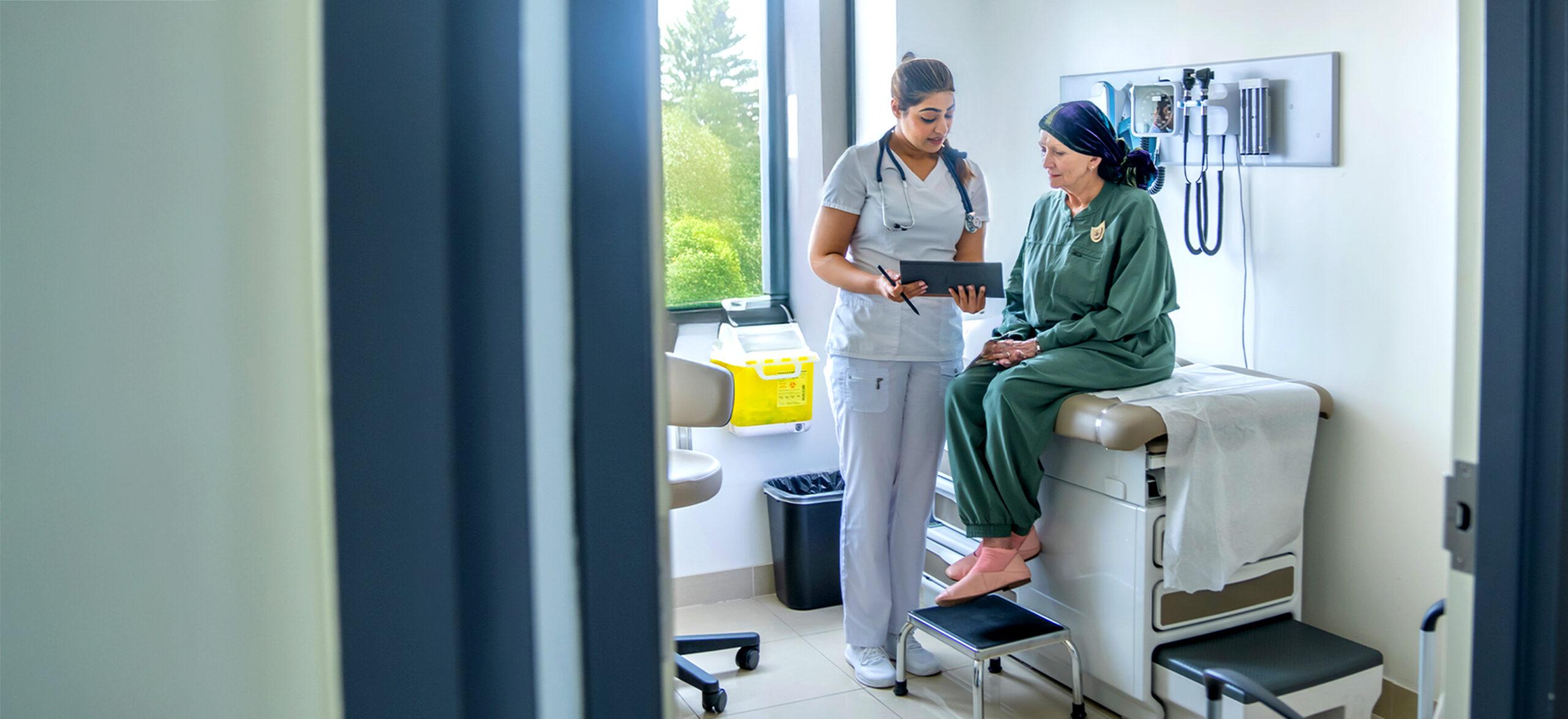 A senior woman who is battling cancer, sits up on an exam table during an appointment. She is dressed comfortably and wearing a headscarf to keep her warm as she talks with the doctor.